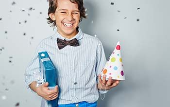 A boy holding a birthday present and birthday hat laughing while confetti is falling in the air