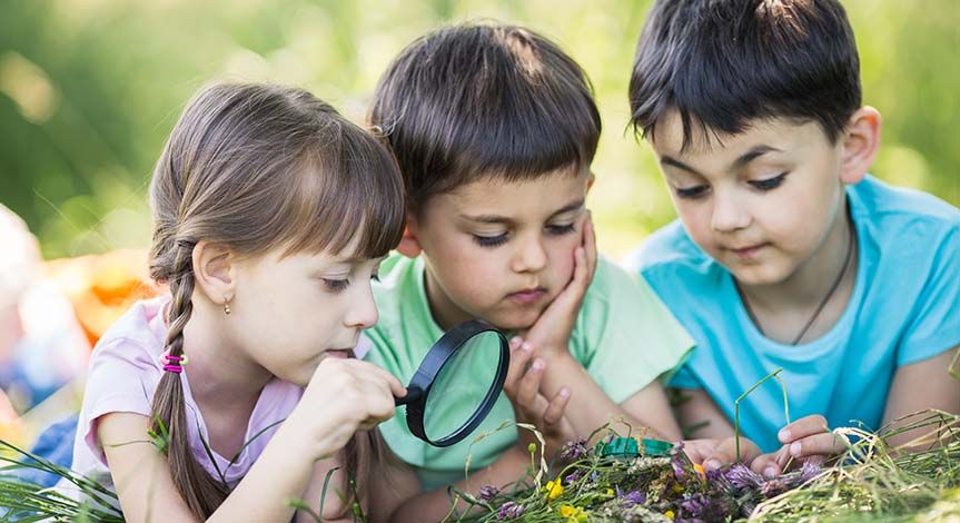 2 boys a girl lying on the grass looking through a magnifying glass