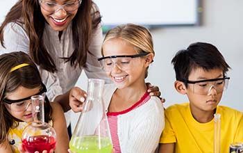 2 girls holding beakers and a boy looking down while teacher points to one of the beakers.