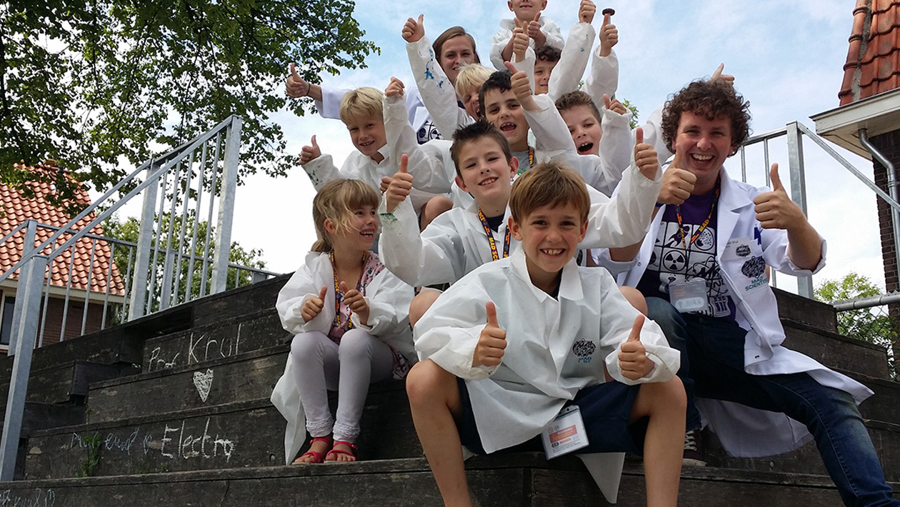 Kids wearing mad science lab coats sitting outside with a mad scientist with their thumbs up