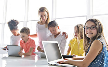 kids with tablets and laptop computer in classroom while teacher stands behind them